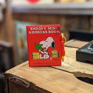 Peut inclure: Un mini carnet d'adresses rouge avec une image de dessin animé de Snoopy en train de lire un livre. La couverture du livre a un cœur rouge. Le carnet d'adresses porte l'inscription "Snoopy Mini Address Book" sur le devant. Le texte "Copr. C1988 United Feature Syndicate, Inc." se trouve en bas du carnet d'adresses.