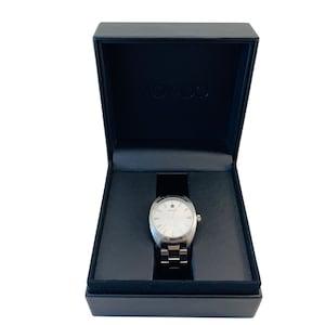 May include: A black watch box with a silver watch inside. The watch has a white face and silver hands.