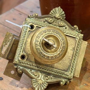 May include: A brass rim lock featuring an ornate rectangular plate with decorative scrollwork and shell motifs at the top and bottom. A central circular knob includes a keyhole with a small metal key inserted. A white plastic zip tie is attached.