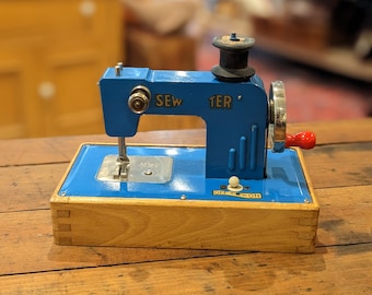 May include: A vintage blue toy sewing machine with the word "SEW TER" printed on the side. The machine has a wooden base and a red handle. The on/off switch is visible. The machine is sitting on a wooden table.