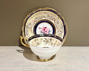 May include: A vintage teacup and saucer set. The white porcelain cup and saucer feature gold floral patterns, a navy blue border, and a pink rose in the center. The cup has a gold handle and base.