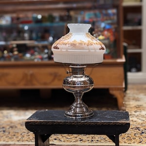 Op de afbeelding: Een zilveren olielamp met een witte glazen kap met een bruin landschapsontwerp staat op een zwarte houten kruk. De lamp staat op een houten vloer met een tapijt met een patroon.