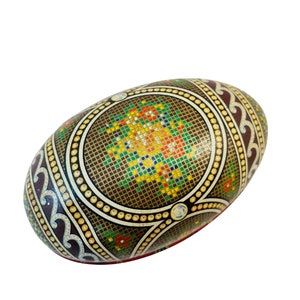 May include: A brown, oval-shaped egg with a colourful geometric design. The egg is decorated with a mosaic of small squares in shades of red, yellow, green, and blue. The design is surrounded by a border of white dots and a band of brown and white swirls.