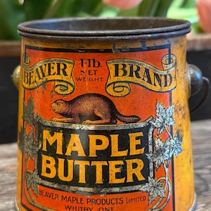 May include: A vintage cylindrical metal tin for Beaver Brand Maple Butter with a 1 lb. net weight label. The orange and black container features a graphic of a beaver, maple leaf illustrations, and text reading Beaver Maple Products Limited.
