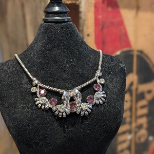 May include: A silver necklace with a delicate chain and a pendant featuring a stylised design with purple and clear rhinestones.
