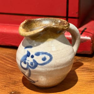 Puede incluir: Jarra de cerámica pequeña, hecha a mano, con cuerpo redondeado y asa. La jarra es blanquecina con borde marrón y diseño floral azul. La jarra mide aproximadamente 7.6 cm de altura.