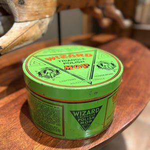 May include: A vintage green Wizard Triangle Polish Mop tin. The tin features the product name in a triangle, with the words "Triangle Polish Mop" and other text. The tin is round and sits on a wooden surface.