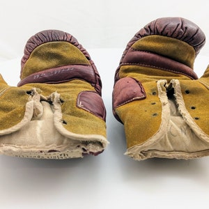 Puede incluir: Un par de guantes de boxeo vintage de cuero marr&oacute;n y beige con un aspecto desgastado y deteriorado. Los guantes tienen un forro blanco y faltan algunas costuras.