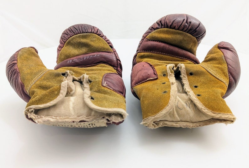 Puede incluir: Un par de guantes de boxeo vintage de cuero marr&oacute;n y beige con un aspecto desgastado y deteriorado. Los guantes tienen un forro blanco y faltan algunas costuras.