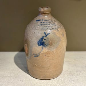 May include: A stoneware jug with a rounded body and a narrow neck. The jug is tan with blue painted details, including a stylized bird and text. The jug is likely an antique or vintage piece.