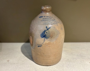 Puede incluir: Jarra de gres beige con detalles pintados en azul, incluyendo un motivo de pájaro y texto. La jarra tiene un cuerpo redondeado y un cuello estrecho. Probablemente un artículo antiguo o vintage.