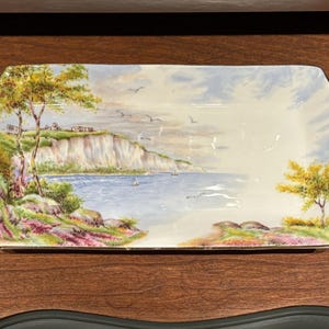 May include: A white porcelain serving tray with gold trim features a painted landscape scene of a coastline with cliffs, trees, and sailboats. The tray has two handles.