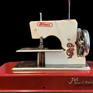 May include: A vintage Straco Jet Sew-O-Matic sewing machine featuring a cream-colored body with red decorative scrollwork and a red metal base. The machine includes a silver hand wheel, a needle assembly, and the text Straco and Jet Sew-O-Matic.