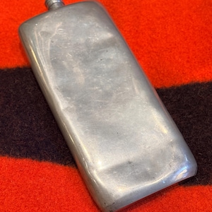 Puede incluir: Una petaca rectangular de color plateado con un cuello pequeño y roscado. La petaca tiene bordes redondeados y una superficie ligeramente texturizada. Descansa sobre un fondo de tela a rayas rojas y negras.