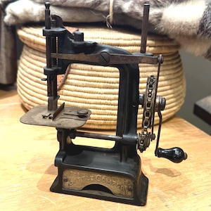 May include: Vintage black cast iron sewing machine with a hand crank and the words "THE LITTLE COMFORT" on a brass plate. The machine is on a wooden surface, with a woven basket and fur in the background.