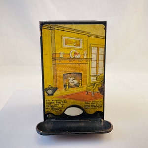 Op de afbeelding: Vintage metalen luciferdooshouder met een zwarte afwerking en een decoratief beeld van een open haard met een geel en baksteenontwerp. De tekst aan de voorkant luidt "Safe - Murrey Mfg. Co. - Jacques, Bary & Co. - Coal, Coke and Charcoal - Office 10 Beaver St. - New York City - Made in USA".