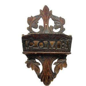 May include: A dark brown wooden wall-mounted comb holder with intricate carvings. The holder has a shelf with the word "COMB" carved into it.