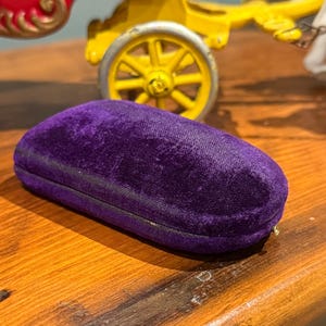 以下が含まれることがあります： 濃い紫色のベルベットの眼鏡ケース。ケースは楕円形で、ジッパー開閉と金色のジッパープルが付いています。ケースは木製の表面に置かれており、おもちゃのワゴンの背景がぼやけています。