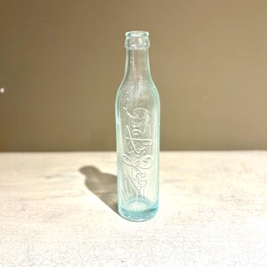 May include: A clear, vintage-style glass bottle with embossed lettering. The bottle has a light blue tint and a slender shape, with vertical lines running down its sides. The word "Coca-Cola" is visible on the bottle.