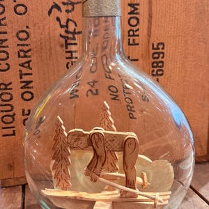 May include: A clear glass bottle containing a miniature wooden diorama of a person skiing among pine trees. The bottle features a beige cap and printed text including "LIQUOR CONTROL," "ONTARIO, OTTAWA," "24 OZ," and "NO FROST." The glass is transparent.