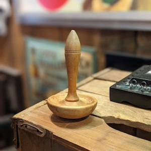 Op de afbeelding: Een houten spoel met een puntige bovenkant en een brede basis. De spoel ligt op een houten oppervlak.