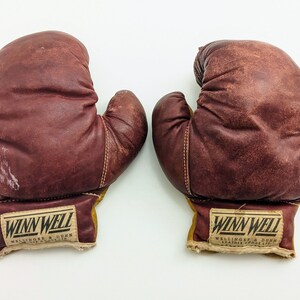 Puede incluir: Un par de guantes de boxeo de cuero marr&oacute;n con etiquetas amarillas y blancas que dicen "Winn Well Wellinger & Dunn Leather Goods Co. Ltd."