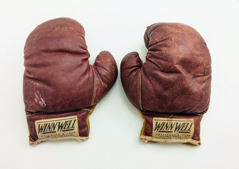 Puede incluir: Un par de guantes de boxeo de cuero marr&oacute;n con etiquetas amarillas y blancas que dicen "Winn Well Wellinger & Dunn Leather Goods Co. Ltd."