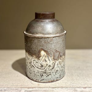 以下が含まれることがあります： ドーム型の蓋が付いたヴィンテージの金属製キャニスター。キャニスターはシルバーで、茶色の蓋と装飾的なエッチングデザインが施されています。エッチングデザインは、花柄と幾何学模様が特徴です。キャニスターは、おそらく保管に使用されます。