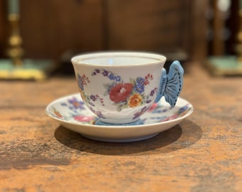 May include: A white teacup with a floral pattern and a blue butterfly handle. The teacup is sitting on a matching saucer.