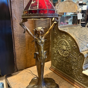 May include: An antique bronze table lamp with a red stained glass shade. The lamp features a female figure holding the shade aloft. The base of the lamp has a floral design. The lamp is on a wooden surface.