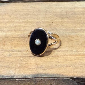Peut inclure: Bague en or avec une pierre centrale ovale noire et une petite perle blanche ronde au centre. La bague a un design &agrave; tige fendue et est pos&eacute;e sur une surface en bois.
