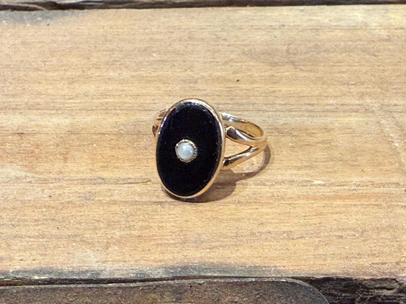Peut inclure: Bague en or avec une pierre centrale ovale noire et une petite perle blanche ronde au centre. La bague a un design &agrave; tige fendue et est pos&eacute;e sur une surface en bois.
