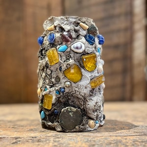 Peut inclure: Un vase en céramique blanche décoré d'une mosaïque de pièces de verre et de métal colorées. Le vase a une forme cylindrique et mesure environ 15 cm de haut. La mosaïque comprend des pièces jaunes, bleues, turquoise et blanches.