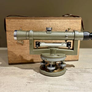 May include: A vintage green metal theodolite surveying instrument with a wooden carrying case. The theodolite has a telescope, a level, and a tripod.