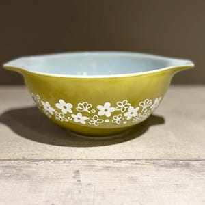 Könnte beinhalten: Eine Vintage Pyrex Rührschüssel in Olivgrün, verziert mit einem weißen Blumenmuster. Die Schüssel hat eine hellblaue Innenseite und zwei Griffe. Dieser Küchenartikel ist perfekt zum Mischen und Servieren.
