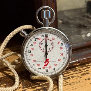 May include: A vintage stopwatch with a white face, black and red numerals, and a silver metal casing. The watch has a cream-colored cord and the text "SWISS MADE". The stopwatch is likely used for timing events.