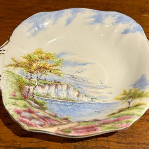 May include: A white ceramic bowl with a blue and green landscape scene. The bowl has a scalloped edge and gold trim.