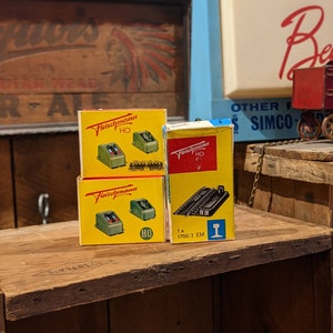 May include: Three boxes of Fleischmann HO scale model train accessories. The boxes are yellow with red and black lettering. The boxes contain a switch, a turnout, and a track section. The boxes are on a wooden surface.