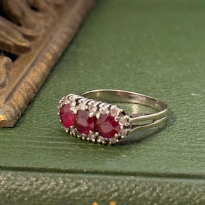 May include: A silver ring featuring three oval, deep red gemstones, likely rubies, set in a row. The ring is accented with smaller, clear stones, possibly diamonds, and rests on a green surface. Jewelry.