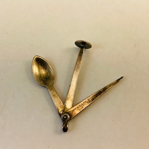 Może przedstawiać: Srebrny wielofunkcyjny przyrząd z łyżką, małym okrągłym narzędziem i ostrym narzędziem. Narzędzia są zawiasowe i składają się razem.