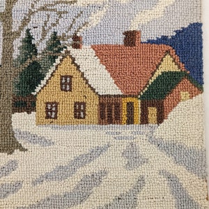 Op de afbeelding: Een handgestikt tapijt dat een wintersc&egrave;ne voorstelt met een klein geel huis, een boom en een met sneeuw bedekte grond. Het huis heeft een rood dak en een schoorsteen waar rook uit komt.
