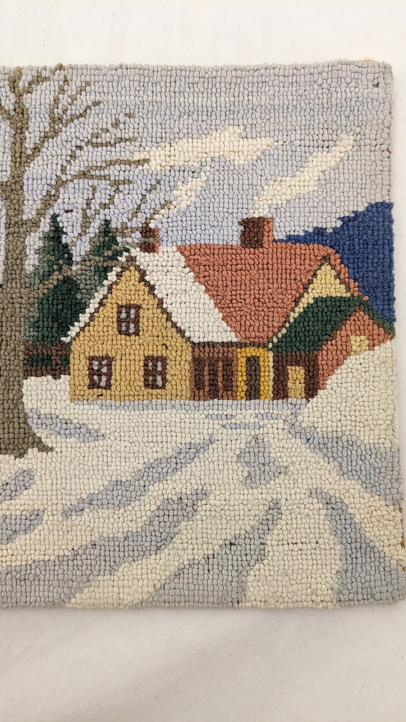 Op de afbeelding: Een handgestikt tapijt dat een wintersc&egrave;ne voorstelt met een klein geel huis, een boom en een met sneeuw bedekte grond. Het huis heeft een rood dak en een schoorsteen waar rook uit komt.