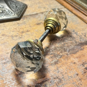 Op de afbeelding: Een heldere, facetgeslepen glazen deurknop met messing accenten. De knop heeft een diamantachtige vorm en is bevestigd aan een metalen staaf. De messing hardware vertoont tekenen van ouderdom. De knop ligt op een houten oppervlak.