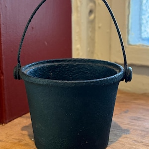 May include: A small, black, cast iron bucket with a textured, matte finish. The bucket features a rounded, thin metal handle attached to two circular side mounts. The vessel has a wide, open top and a tapered body with a flat, circular base.