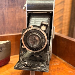 May include: Vintage black and silver folding camera with a lens marked "Compur" and "2. Deckel-München." The camera is open, revealing the bellows and lens. The camera is on a wooden surface.