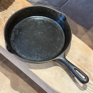 Op de afbeelding: Een goed ingebakken, zwarte gietijzeren koekenpan met een lange handgreep en een ophanglus. De pan staat op een lichtgekleurd houten oppervlak. De koekenpan is rond met een licht verhoogde rand.