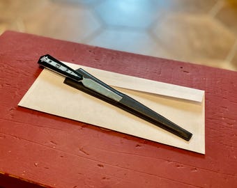Könnte beinhalten: Ein schwarzer Brieföffner mit einer silbernen Klinge und einem schwarzen Ledergriff. Der Griff ist mit einem dekorativen Gebäudemotiv verziert.