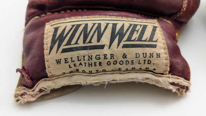Puede incluir: Primer plano de un guante de cuero marr&oacute;n con una etiqueta blanca que dice "Winn Well Wellinger & Dunn Leather Goods Ltd. Toronto Canada".
