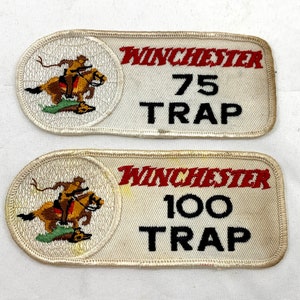 Può includere: Due toppe ricamate bianche con un logo Winchester rosso e nero. La toppa superiore dice "75 Trap" e la toppa inferiore dice "100 Trap". Entrambe le toppe mostrano un cowboy a cavallo.