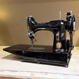 以下が含まれることがあります： 光沢のある黒いSinger 222Kミシン。金属製の針棒、右側のハンドホイール、前面の金色の円形エンブレムが特徴です。このミシンは平らなベースと水平なアーム構造を備えています。